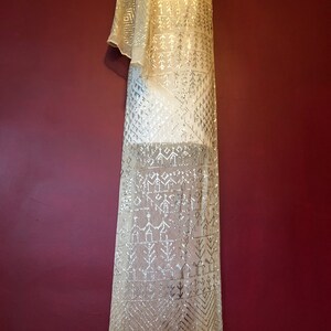 Pu&ograve; includere: Un lungo scialle beige trasparente con un motivo geometrico di paillettes argentate. Lo scialle &egrave; drappeggiato su un manichino.