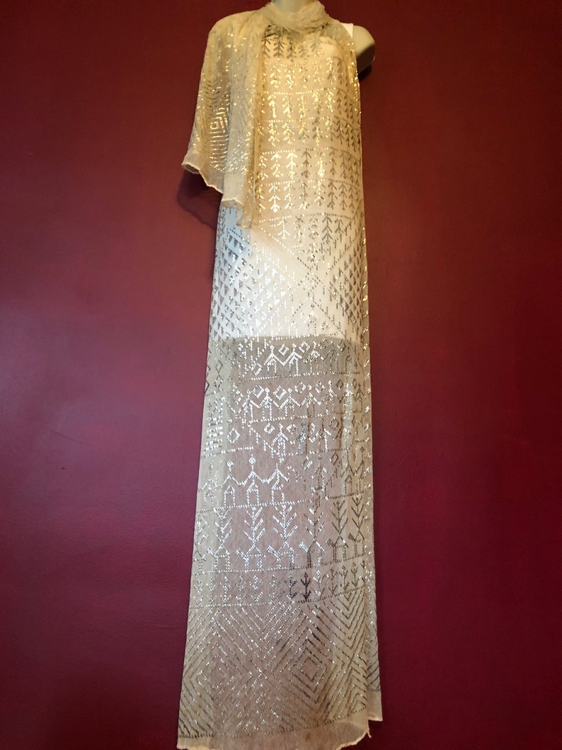 Pu&ograve; includere: Un lungo scialle beige trasparente con un motivo geometrico di paillettes argentate. Lo scialle &egrave; drappeggiato su un manichino.