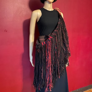 May include: A black shoulder bag with long fringe and red accents. The bag is draped over a mannequin wearing a black top and skirt. The bag's woven fabric has a geometric pattern.