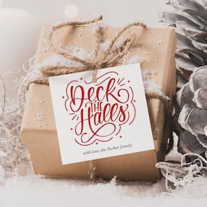 May include: A brown gift box wrapped in twine with a white gift tag that says "Deck the Halls" in red lettering. The gift box is surrounded by white candles, white pine cones, and white faux snow.