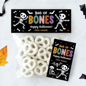 May include: A clear bag of white pretzel twists, with a black label saying "BAG OF BONES" and "Happy Halloween!" featuring skeleton illustrations. A colourful autumn leaf and paper bats are also present.