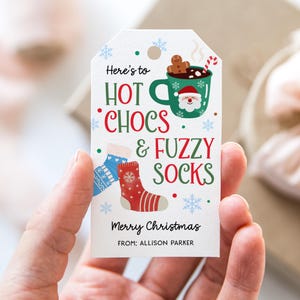 May include: A white gift tag with the text "Here's to HOT CHOCS & FUZZY SOCKS" in green and red. The tag features illustrations of a mug with Santa, socks, and snowflakes. The words "Merry Christmas" are also on the tag.