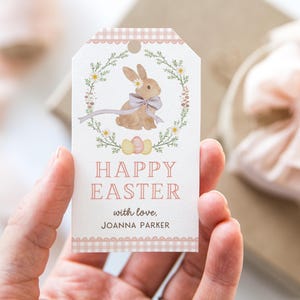 Puede incluir: Una etiqueta de regalo de Pascua decorativa con una ilustración de acuarela de un conejito dentro de una corona floral. La etiqueta dice "Happy Easter" y "with love, Joanna Parker" en texto rosa. Tiene un borde de cuadros.