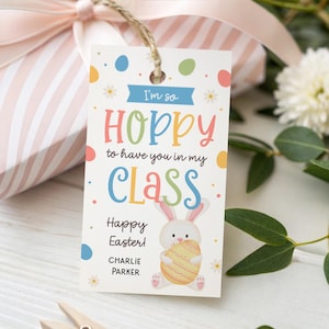May include: A gift tag with the text "I'm so hoppy to have you in my class" and "Happy Easter!" features a cartoon bunny holding an Easter egg. The tag is attached to a wrapped gift with a pink bow.