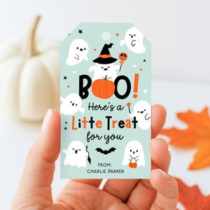 May include: A Halloween-themed gift tag with a light blue background, featuring white ghosts, an orange pumpkin, and the text "BOO! Here's a Little Treat for you." The tag is held in a hand.