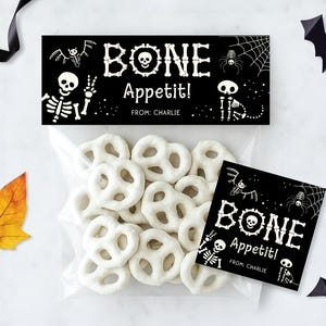 May include: A Halloween-themed treat bag filled with white pretzels. The bag is sealed with a black label that reads "BONE Appetit!" in white, with skeleton and spider illustrations. A smaller matching tag is also present.
