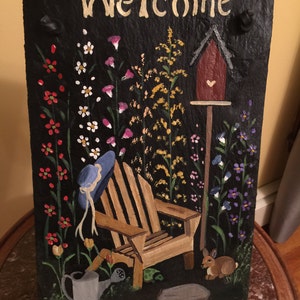 May include: A hand-painted slate welcome sign with a black background. The sign features a whimsical garden scene with a wooden chair, a birdhouse, a watering can, a turtle, a rabbit, and a variety of flowers. The words "Welcome" are painted in white at the top of the sign.