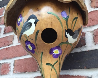 May include: A hand-painted gourd birdhouse with a brown, tan, and green exterior. The birdhouse has a small, round opening for birds to enter. The gourd is decorated with purple flowers and two black and white birds.