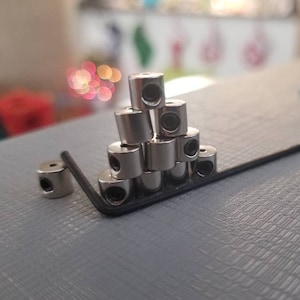 May include: A collection of small, cylindrical metal objects with a hole through the center, stacked in a pyramid shape. A black hex key is positioned near the objects. One object is isolated to the right. The objects appear to be hardware components.