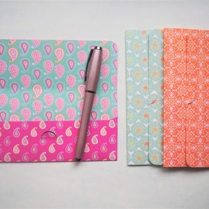 May include: Three decorative envelopes with a paisley pattern. The envelopes are folded and have a flap on the top. The envelopes are in shades of green, pink, and orange. A silver pen is lying on top of the envelopes.