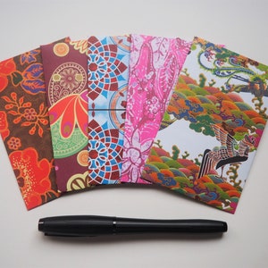 Puede incluir: Seis sobres con patrones coloridos con un bolígrafo negro sobre una superficie blanca. Los sobres presentan varios diseños, incluyendo patrones florales, geométricos y abstractos.