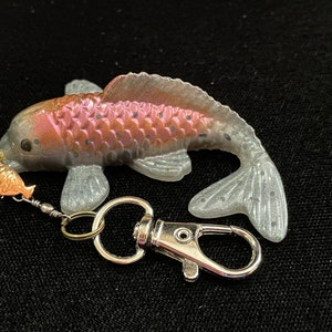 Puede incluir: Un llavero de plata y oro con un dije de pez rosa y blanco. El pez tiene un diseño detallado con escamas y un acabado brillante.