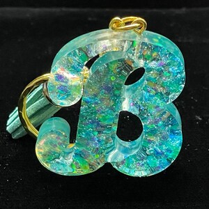 May include: A light blue resin letter B charm with iridescent glitter embedded in the resin. The charm is attached to a gold key ring.