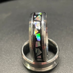 May include: A silver ring with a black inlay and iridescent opal accents. The ring is shown on a black surface.