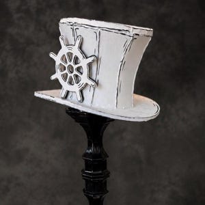 May include: A white top hat with black hand-drawn outlines and a ship's wheel emblem attached to the side. The hat features a flat brim and a tall, slightly curved crown, resting on a black pedestal stand with a decorative, tiered base.
