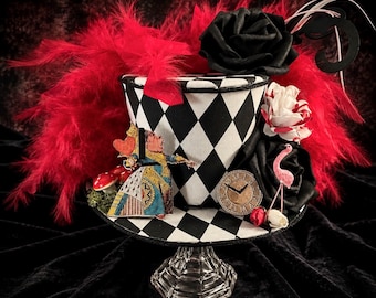 May include: A top hat featuring a black and white diamond pattern, adorned with red feathers, black and white fabric roses, a miniature figure of a queen holding a heart, a small clock face, a pink flamingo figurine, and red mushrooms.