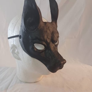 May include: A black dog mask with pointed ears and a detailed snout. The mask is made of a dark material and has eye openings. A black elastic band is attached to the mask for wearing.