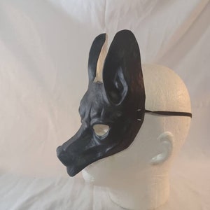 May include: A black leather mask in the shape of a dog or wolf head, with large upright ears and a long snout. The mask is worn on a white mannequin head, with a thin black elastic strap.