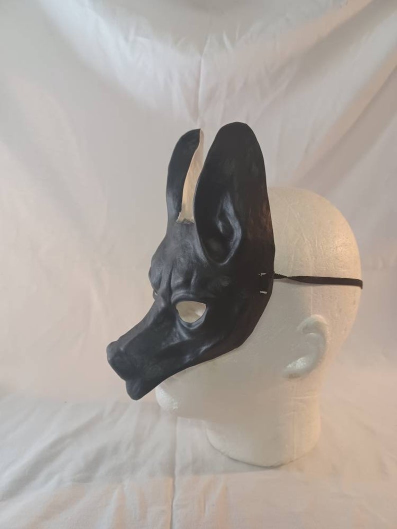 May include: A black leather mask in the shape of a dog or wolf head, with large upright ears and a long snout. The mask is worn on a white mannequin head, with a thin black elastic strap.