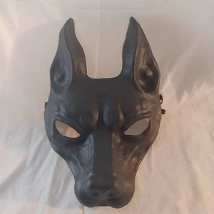 May include: A black dog-shaped mask with large, upright ears and eye openings. The mask is made of a matte material and has a strap on each side for wearing. The mask is designed to be worn on the face.