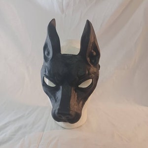 May include: A black mask shaped like a dog's head, with long, pointed ears and a prominent snout. The mask has white eye openings and is displayed on a white mannequin head. The mask is likely for costume or decorative purposes.