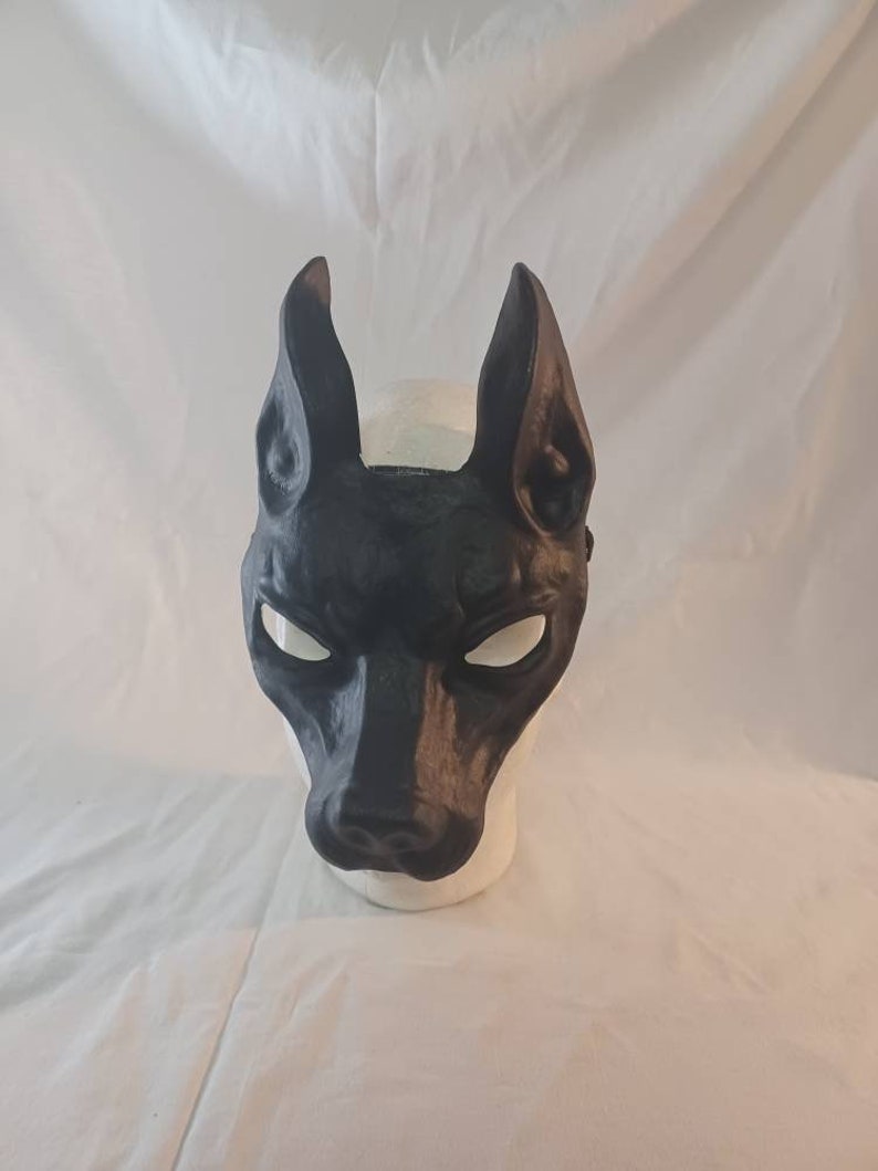 May include: A black mask shaped like a dog's head, with long, pointed ears and a prominent snout. The mask has white eye openings and is displayed on a white mannequin head. The mask is likely for costume or decorative purposes.
