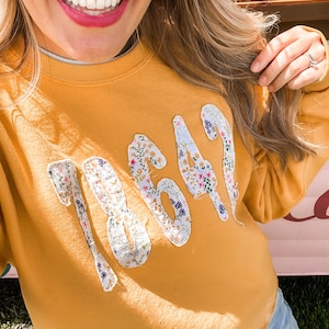 Może przedstawiać: Musztardowa bluza z numerami "18649" naszytymi z przodu. Numery wykonane są z kwiatowej tkaniny z różnorodnymi kolorowymi kwiatami. Bluza ma długie rękawy i okrągły dekolt.