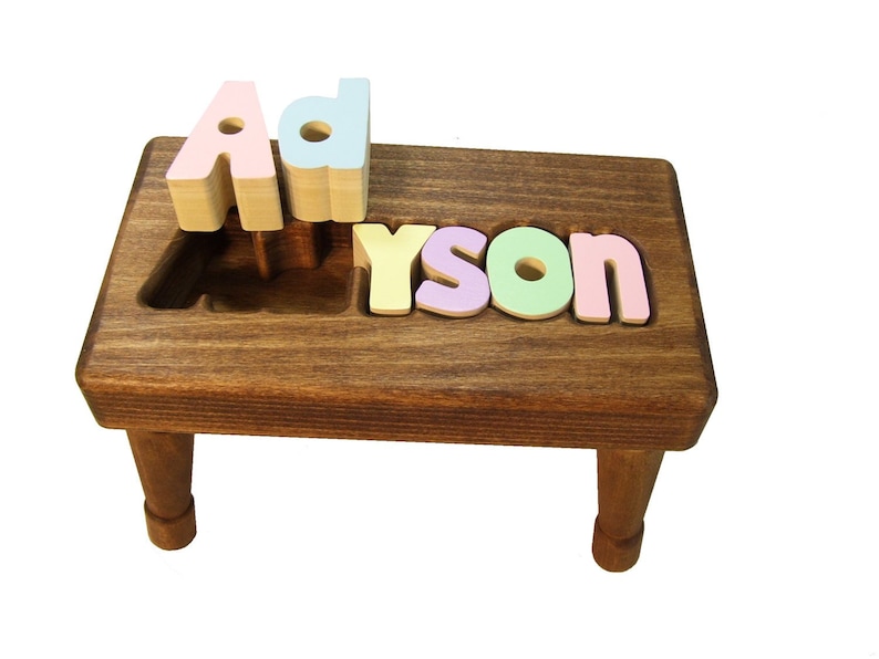 Op de afbeelding: Een houten opstapje met de naam "Ayson" gespeld in kleurrijke houten letters. De letters zijn afneembaar en kunnen als puzzel worden gebruikt.