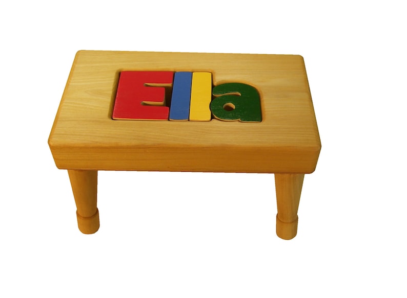 Op de afbeelding: Een houten opstapje met de naam "Ela" gespeld in kleurrijke letters. De letters zijn rood, blauw, geel en groen.