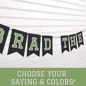 May include: Black and green banner with the text "Brad the Grad" hanging on a white wall. The banner is made of felt and has a vintage look.