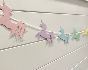 May include: A pastel rainbow unicorn banner with six unicorns in different pastel colors, including pink, lavender, light blue, green, yellow, and orange. The unicorns are strung together with white string.