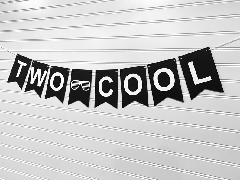May include: Black banner with white text spelling "TWO COOL" and a sunglasses graphic. The banner is strung on a white string and hangs against a white, horizontally-lined wall. Party decoration.