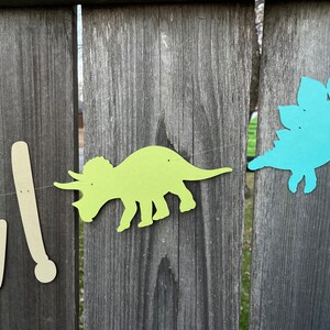 May include: A green dinosaur cutout with a blue dinosaur cutout on a wooden fence. The dinosaurs are part of a banner that reads "One!"