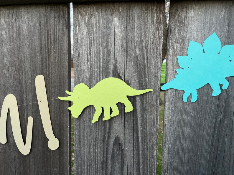 May include: A green dinosaur cutout with a blue dinosaur cutout on a wooden fence. The dinosaurs are part of a banner that reads "One!"