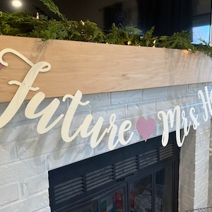 May include: White wooden letters spelling "Future Mrs. Hart" with pink hearts in between the words. The letters are hanging on a wooden mantel above a white brick fireplace.