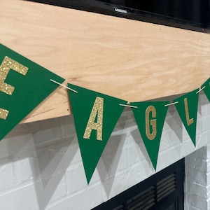 May include: Green and gold felt pennant banner with the word "EAGLES" in gold glitter letters. The banner is strung up on a wall.