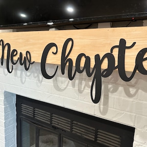 May include: A black wooden sign with the words "A New Chapter" in a cursive font. The sign is mounted above a white brick fireplace.