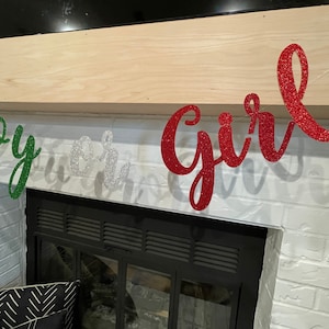 May include: A gender reveal banner with the words "Boy or Girl" in green, silver, and red glitter letters. The banner is hanging above a fireplace.