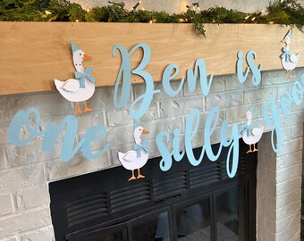 Könnte beinhalten: Ein hellblaues Banner mit den Worten "Ben is one silly goose" in Schreibschrift, verziert mit Cartoon-Gänsen. Das Banner wird über einem Kamin mit einem hölzernen Kaminsims und weißen Ziegeln angezeigt.
