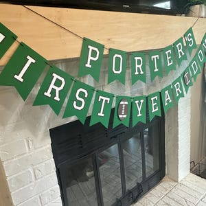 May include: A green banner with white lettering that reads "PORTER'S FIRST YEAR DOWN" is strung above a fireplace. A football graphic is between the words "YEAR" and "DOWN". The fireplace is white brick with a black firebox.