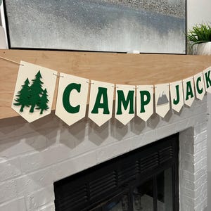 May include: A banner with the words "CAMP JACK" in green letters. The banner is made of off-white pennants. The left side features green tree cutouts, and a mountain graphic is on the right. The banner is hanging above a fireplace.