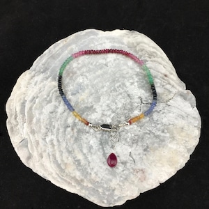 Puede incluir: Un delicado brazalete con una gama de gemas coloridas. El brazalete está compuesto por pequeñas cuentas redondas en tonos rojo, rosa, verde, negro, azul y amarillo. Un cierre plateado y un colgante de rubí completan el diseño. El brazalete se exhibe sobre una superficie blanca texturizada.