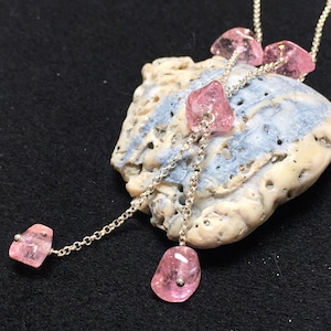 Op de afbeelding: Een zilveren ketting met roze edelsteen hangers. De ketting wordt getoond op een schelp.