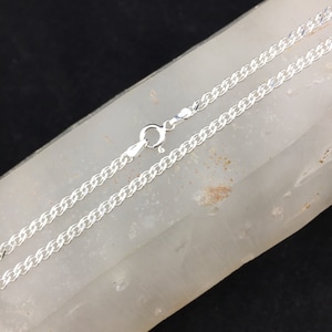 May include: A silver curb chain necklace with a lobster clasp closure. The chain is laid out on a white surface.