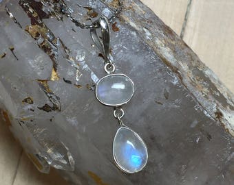 May include: A silver chain necklace with a teardrop-shaped moonstone pendant and a smaller oval moonstone pendant. The necklace is displayed on a clear quartz crystal.