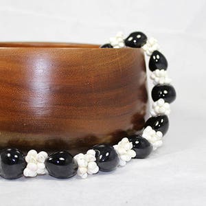 May include: A black and white lei made of small, round beads. The lei is draped over a wooden bowl.