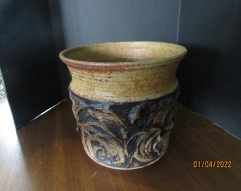 May include: A brown and tan ceramic planter with a decorative design of stylized leaves and swirls. The planter has a wide rim and a slightly flared base.