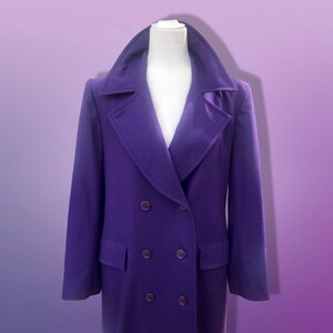 May include: A double-breasted purple coat with a wide collar and two front pockets. The coat is displayed on a white mannequin against a purple gradient background. The coat has six buttons.