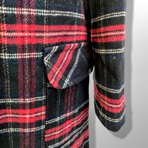 May include: Close-up of a black, red, and white plaid coat. The coat features a pocket on the front and a long sleeve. The plaid pattern includes red, white, and blue stripes. The coat is made of a thick, textured fabric.