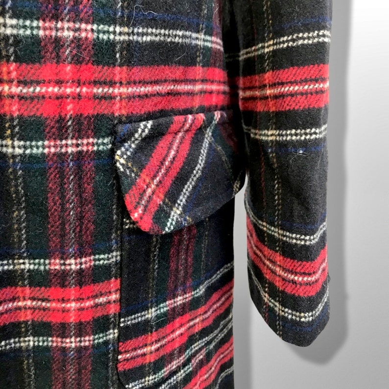 May include: Close-up of a black, red, and white plaid coat. The coat features a pocket on the front and a long sleeve. The plaid pattern includes red, white, and blue stripes. The coat is made of a thick, textured fabric.